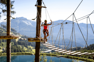 Frau klettert im Hochseilgarten. Hinter ihr sieht man die Bergkulisse mit See.