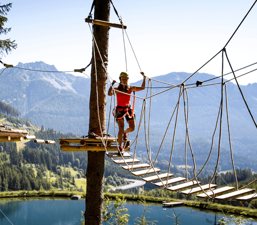 Frau klettert im Hochseilgarten. Hinter ihr sieht man die Bergkulisse mit See.