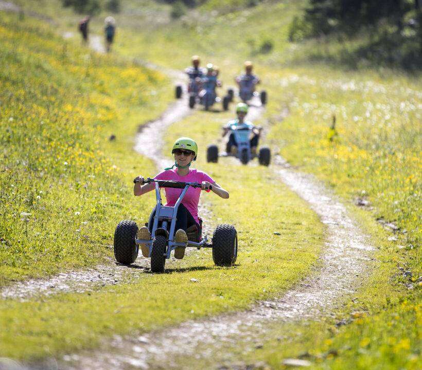 Eine Gruppe von Menschen sitzen auf Mountaincarts und fahren einen Bergweg hinunter.