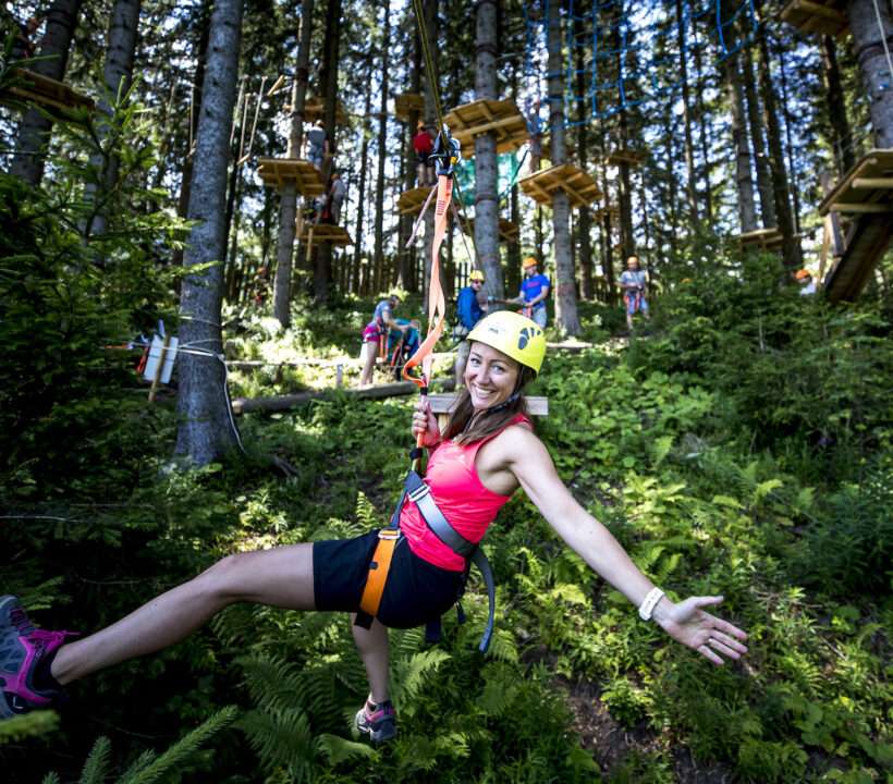 Eine Frau in Klettermontur trägt einen gelben Helm und fliegt im Kletterpark am Präbichl mit der Zipline. Dabei lächelt sie.