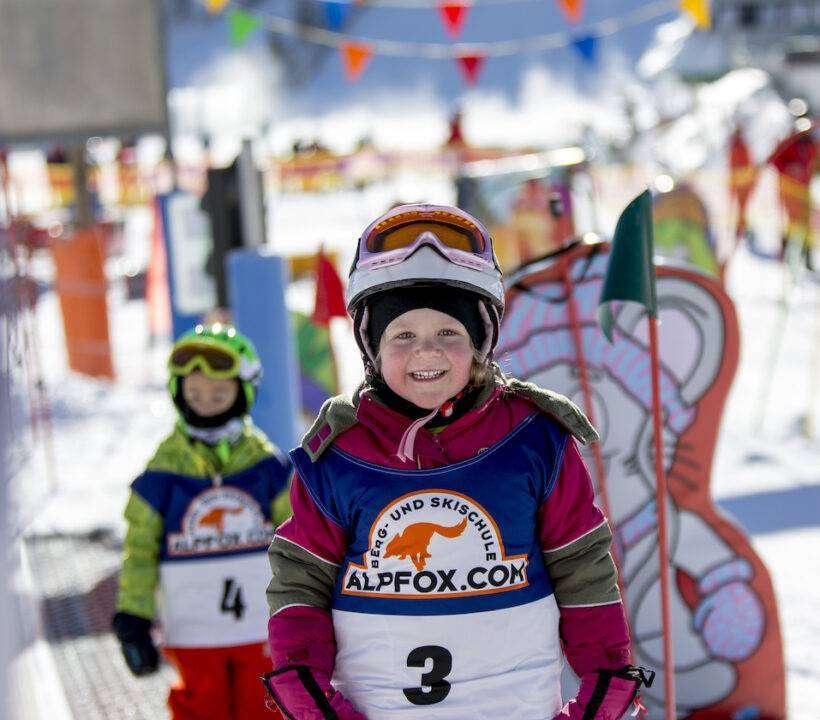 Skikinder stehen vor bunten Skifiguren und Fähnchen und lächeln in die Kamera