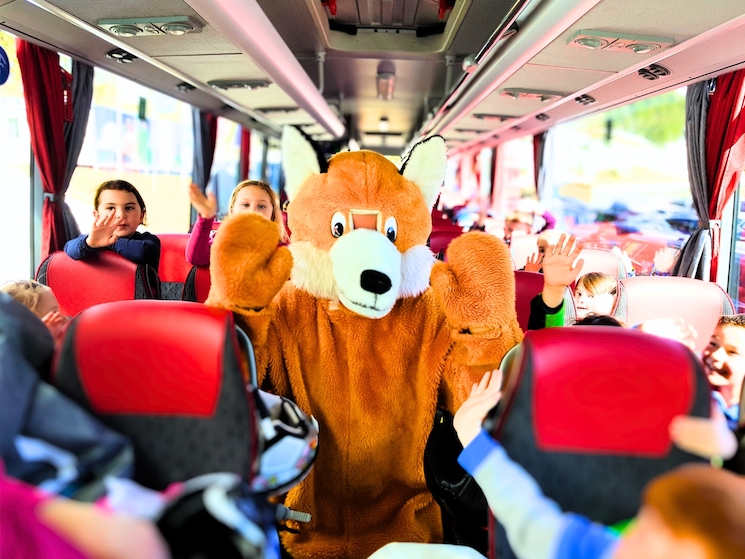 Das Alpfox-Maskottchen "Foxi - der Fuchs" steht im Gang des Skibusses und winkt zusammen mit den Skikindern in die Kamera.