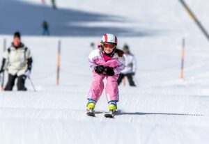 Kind fährt auf Ski auf einer Wellenbahn im Schuss und lächelt in die Kamera.